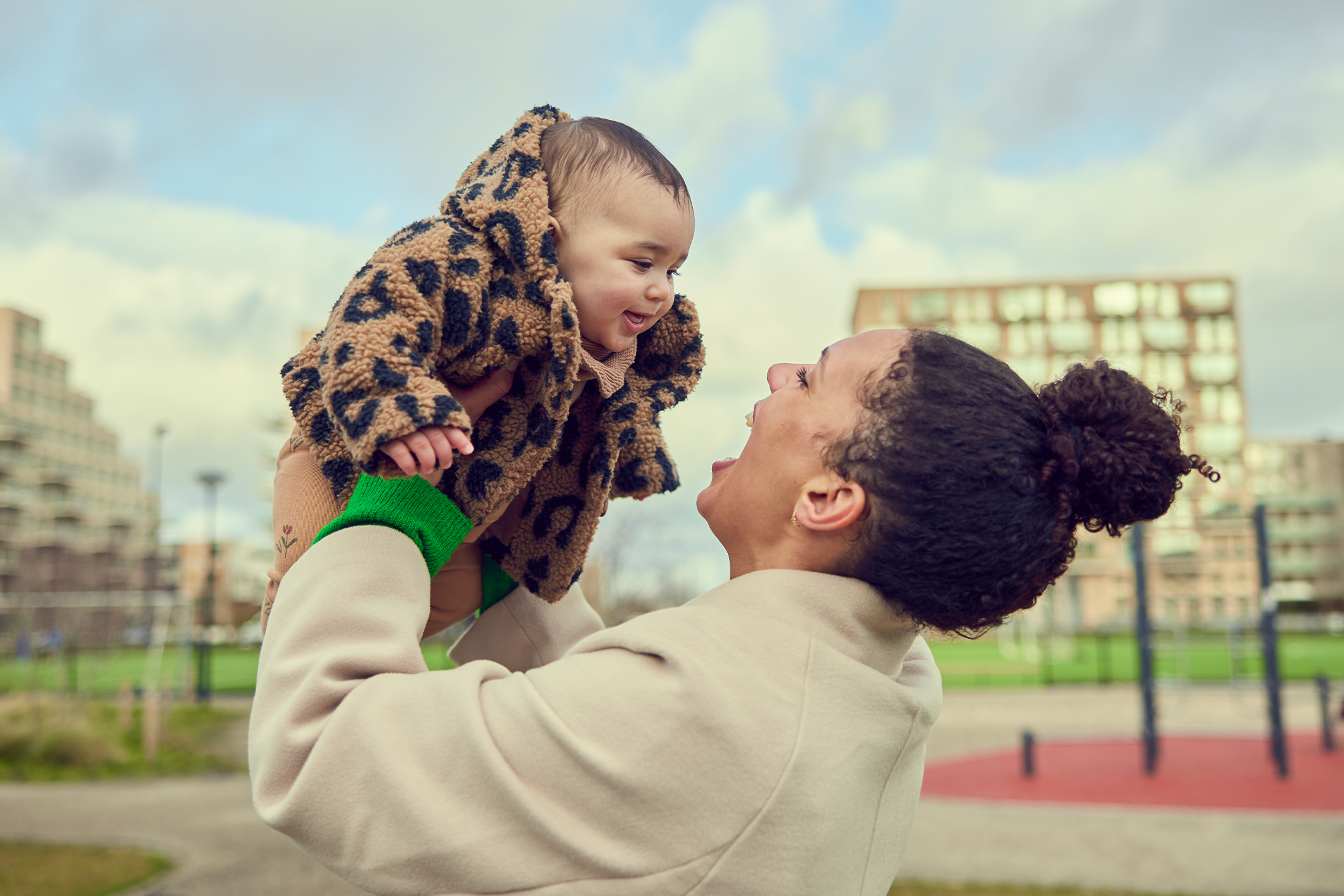 Moeder die een baby omhoog houdt en lacht. Ze zijn in een speeltuin